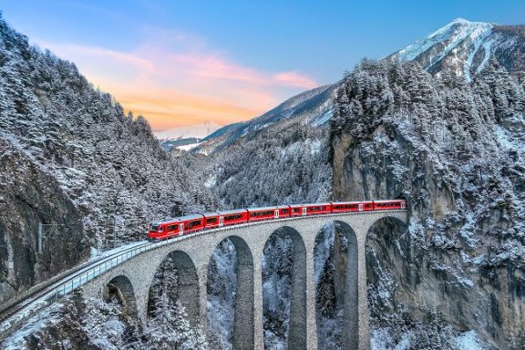 Glacier Express Train rouge traversant un viaduc dans un paysage montagneux enneigé au crépuscule.