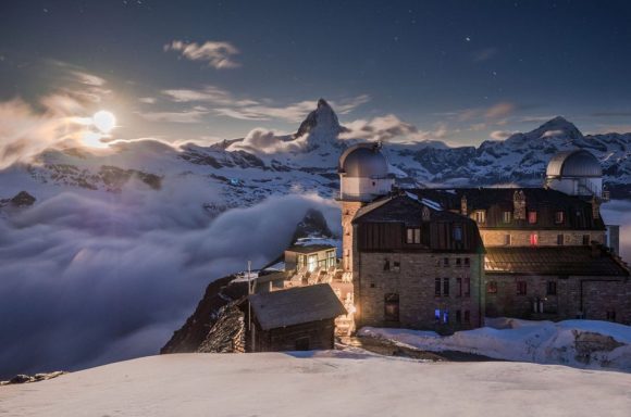 Kulmhotel Gornergrat Montagne enneigée avec un bâtiment éclairé, sous une pleine lune et des nuages.