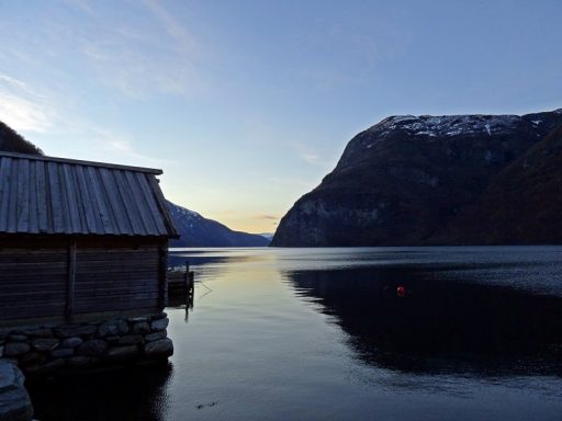 Cabane en bois au bord d'un fjord, montagnes en arrière-plan sous un ciel bleu.